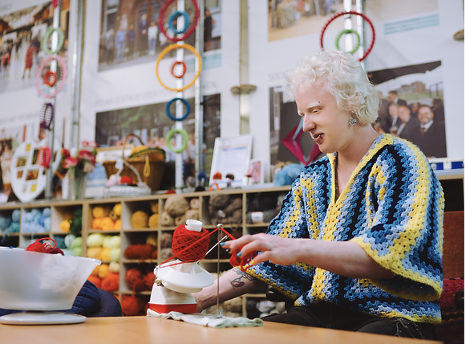 Marnix, person with Albinism is knitting around him there is a lot of balls of yarn. Marnix is winding bright red yarn using a yarn winding around, he wears a knitted sweater in bright colours, yellow and different shades of blue.